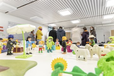 Playmobilfiguren zeigen einen möglichen Aufbau für einen ersten Besuch im Dorf. Dahinter diskutiert eine Gruppe von Teilnehmenden